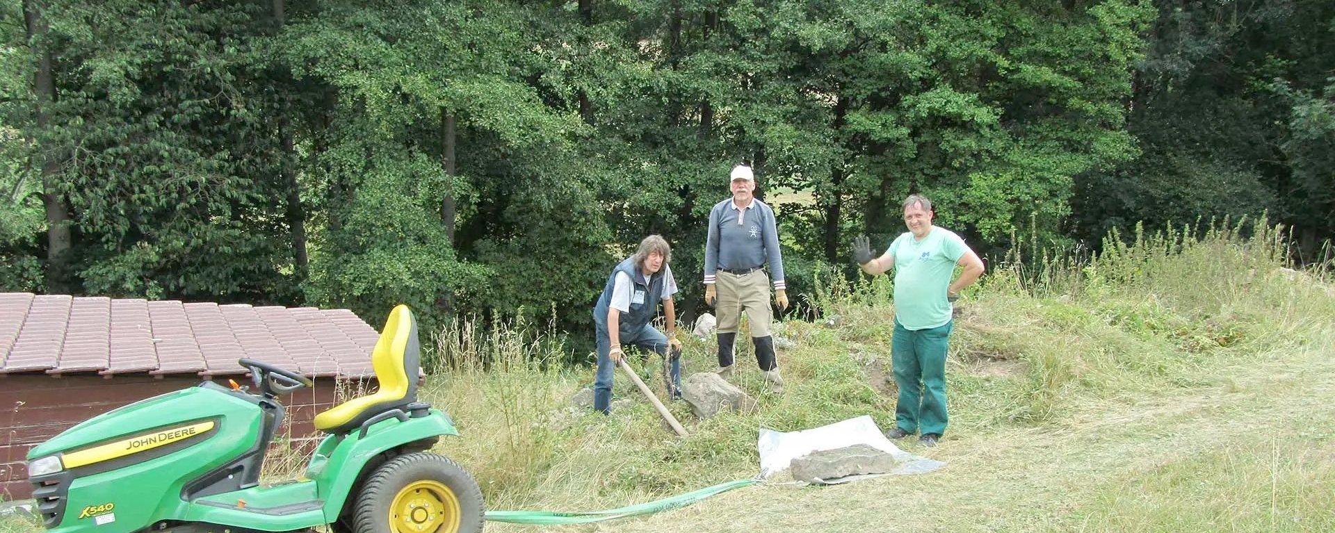 Drei Männer bei der Gartenarbeit auf einer Wiese. Sie tragen Handschuhe oder haben Gartengerät in der Hand - daneben ein Mini-Traktor