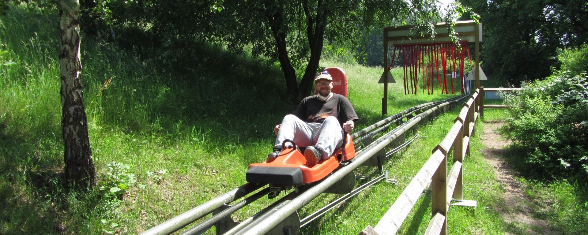 Actionfoto: Ein Mann mittleren Alters mit Bart und Brille sitzt in einem Sommer-Rodel und bremst, während er in die Kamera lächelt