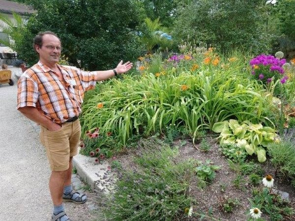 Eine Hand in der Hosentasche: stolz präsentiert ein Mann mittleren Alters und Halbglatze mit ausgestrecktem Arm das Blumen- und Kräuterbeet