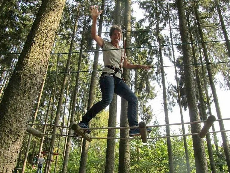 Hochseilgarten: Eine junge Frau balanciert freihändig, in die Kamera winkend über eine art Hängebrücken-Konstruktion