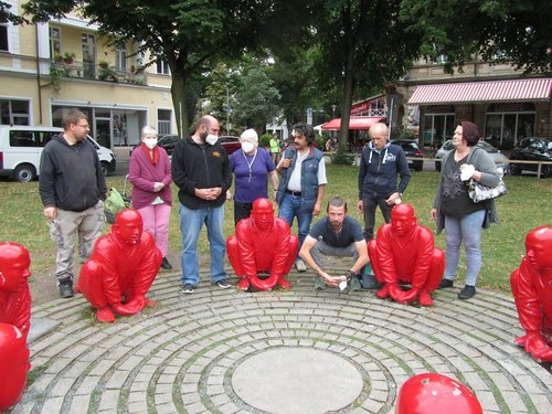 Frauen und Männer stehen um ein Kunstobjekt, dass einzelne rot lackierte Mönche in hockender Position zeigt. Ein Mann hockt sich zu den Mönchen und nimmt die gleiche Pose ein.