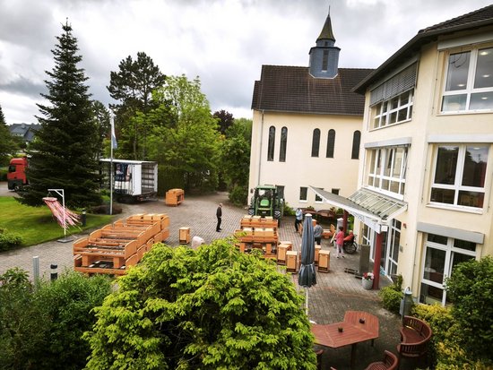 Auf einem gepflasterten Hof vor einem hellen Gebäude mit Turm liegen zahlreiche aufgestapelte Kirchenbänke. Ein grüner Traktor steht in der Mitte. Im Hintergrund parkt ein weißer LKW. Mehrere Personen, darunter einer im Rollstuhl, beobachten die Arbeiten.